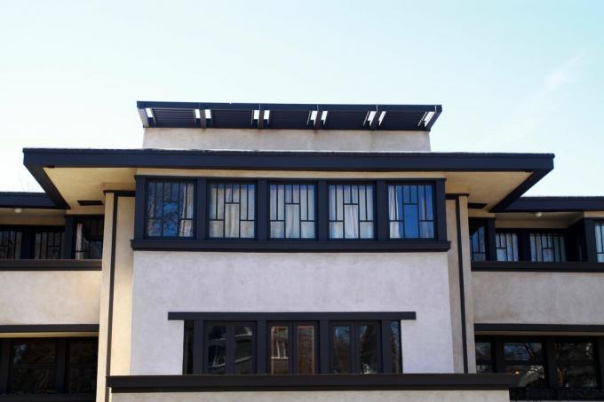 vaakasuoraan suuntautuva vaaleanpuoleinen talo tummanruskealla koristelulla, litteä katto valtaisilla ulokkeilla ja ikkunarivit lyijylasilla